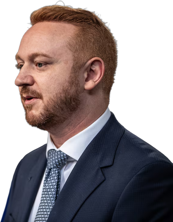 Mickey Amery in focus at the front of the frame, looking away from the camera, with Premier Danielle Smith slightly blurred in the background, both standing indoors at a formal political event.